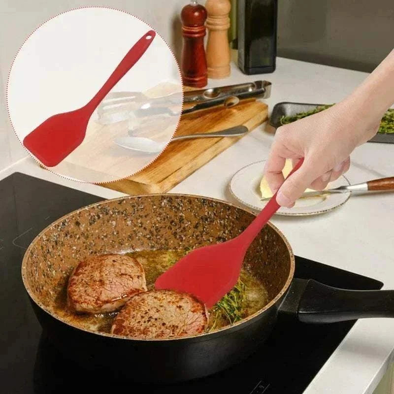 Red Silicone Cooking Spatula with flexible head being used to cook two pieces of meat in a black frying pan on a stovetop